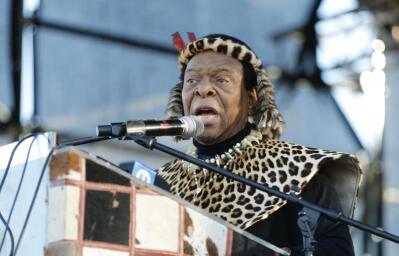 A man in traditional leopardskin headgear and robes behind a microphone and a lectern. 