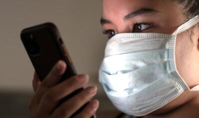 A woman wears a mask and looks at a smartphone.