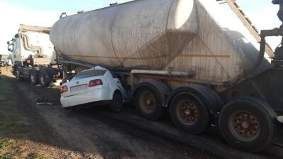 The rear end of a white car crushed underneath a large truck trailer.