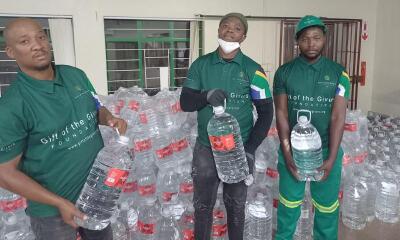 Three Gift of the Givers volunteers each holding a bottle of water. In the background a large number of water bottles for donation. 